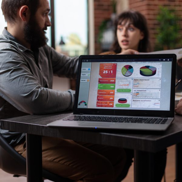Laptop computer having marketing project graphs on screen while multiethnic businesspeople discussing company turnover in startup office. Diverse team planning business collaboration